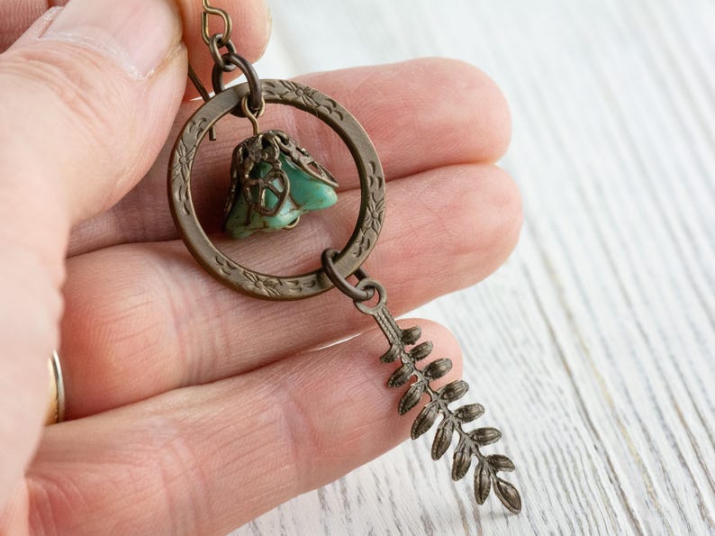 May include: A bronze-coloured earring with a teal stone pendant and a fern-shaped charm. The earring has a circular design with detailed patterns. The earring is held in a person's hand.