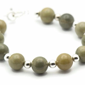 K&ouml;nnte beinhalten: Ein Armband aus gr&uuml;nen und braunen Steinperlen mit silbernen Akzenten. Das Armband besteht aus runden Perlen und hat ein schlichtes Design.