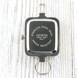 Puede incluir: Reloj Geneva negro y plateado con esfera rectangular, llavero y el texto "STAINLESS STEEL BACK", "GENEVA JAPAN MOVT 2039-T WATER RESISTANT". El reloj est&aacute; sobre una superficie de madera blanca.