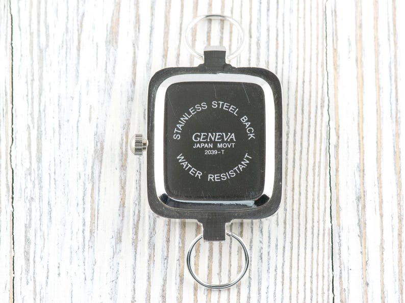 Puede incluir: Reloj Geneva negro y plateado con esfera rectangular, llavero y el texto "STAINLESS STEEL BACK", "GENEVA JAPAN MOVT 2039-T WATER RESISTANT". El reloj est&aacute; sobre una superficie de madera blanca.