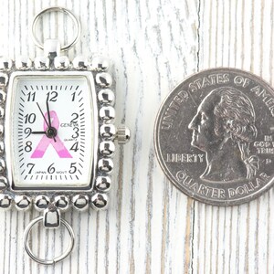 May include: Silver-toned watch with a rectangular face and a pink ribbon design. The watch face displays the time and the word "GENEVA". A U.S. quarter is next to the watch for size comparison.