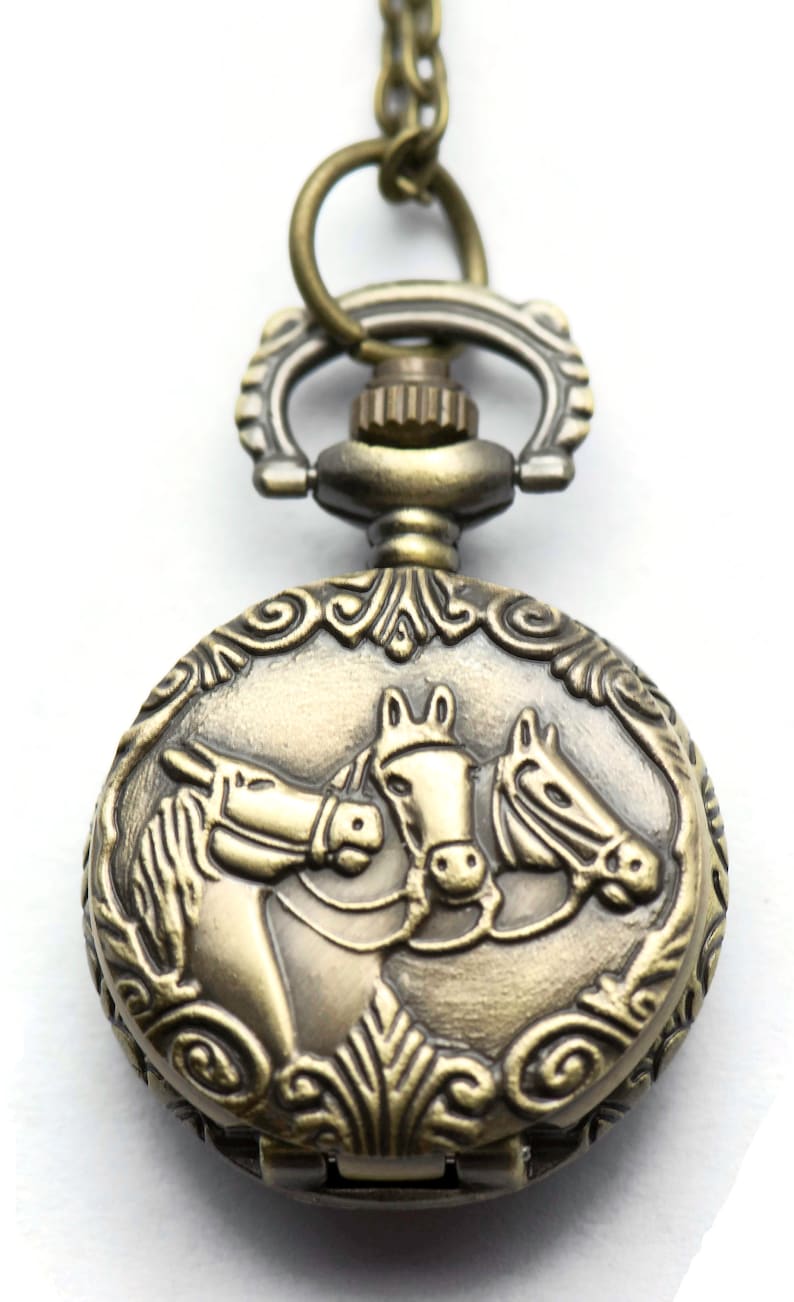 Puede incluir: Un reloj de bolsillo de bronce con una cadena, que presenta un dise&ntilde;o ornamentado y tres cabezas de caballo en la parte delantera.