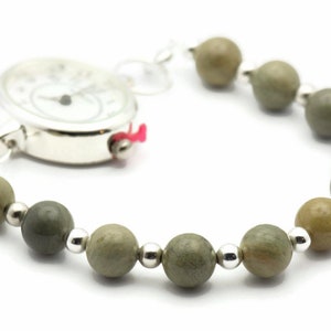 K&ouml;nnte beinhalten: Eine silberne Taschenuhr mit wei&szlig;em Zifferblatt und schwarzen Zeigern ist an einem Armband aus gr&uuml;nen und braunen Perlen befestigt.