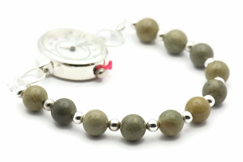 K&ouml;nnte beinhalten: Eine silberne Taschenuhr mit wei&szlig;em Zifferblatt und schwarzen Zeigern ist an einem Armband aus gr&uuml;nen und braunen Perlen befestigt.
