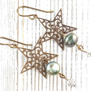 May include: A pair of bronze-colored star-shaped earrings with intricate filigree detailing. Each earring features a gray-green pearl and a clear crystal bead. The earrings hang from bronze-colored ear wires.