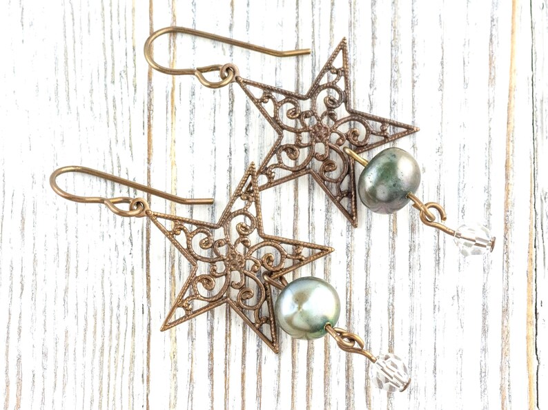 May include: A pair of bronze-colored star-shaped earrings with intricate filigree detailing. Each earring features a gray-green pearl and a clear crystal bead. The earrings hang from bronze-colored ear wires.