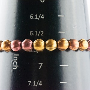 May include: A bracelet made of small, round beads in shades of gold and rose gold. The bracelet is measured on a ruler that shows it is 6 1/2 inches in length.