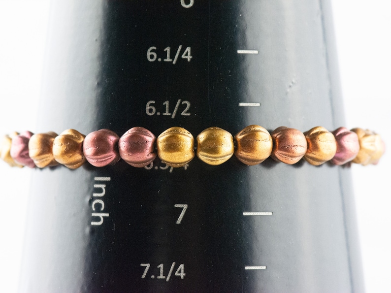 May include: A bracelet made of small, round beads in shades of gold and rose gold. The bracelet is measured on a ruler that shows it is 6 1/2 inches in length.