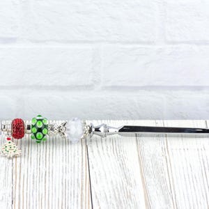 May include: A decorative letter opener with a silver blade and handle. The handle is adorned with colorful beads, including red, green, and white, and a small Christmas tree charm. The opener rests on a white wooden surface.