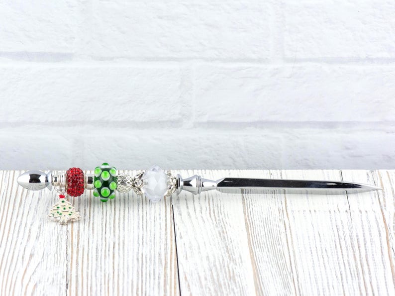 May include: A decorative letter opener with a silver blade and handle. The handle is adorned with colorful beads, including red, green, and white, and a small Christmas tree charm. The opener rests on a white wooden surface.