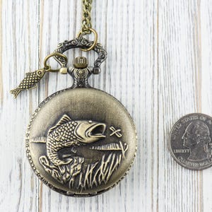 Pode incluir: Um rel&oacute;gio de bolso de bronze com uma corrente e um amuleto de peixe. O mostrador do rel&oacute;gio apresenta uma imagem gravada de um peixe saltando para fora da &aacute;gua. O rel&oacute;gio &eacute; aproximadamente do tamanho de uma moeda de euro.