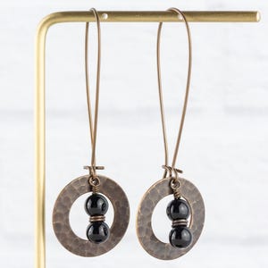 Peut inclure: Une paire de boucles d'oreilles artisanales avec des pendentifs circulaires de couleur cuivre martel&eacute;. Chaque pendentif contient deux perles noires brillantes. Les boucles d'oreilles sont suspendues &agrave; de longs fils fins de couleur cuivre.