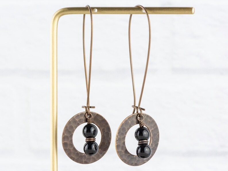 Peut inclure: Une paire de boucles d'oreilles artisanales avec des pendentifs circulaires de couleur cuivre martel&eacute;. Chaque pendentif contient deux perles noires brillantes. Les boucles d'oreilles sont suspendues &agrave; de longs fils fins de couleur cuivre.