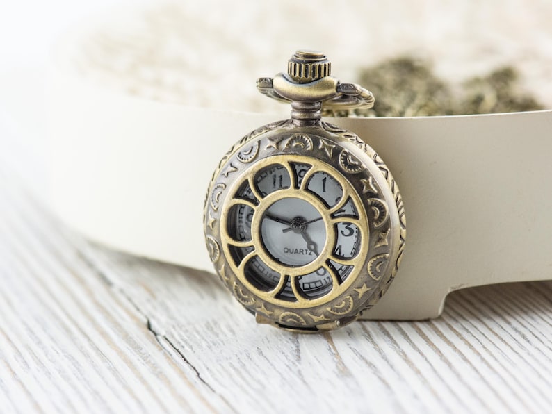 Puede incluir: Un reloj de bolsillo dorado con un dise&ntilde;o ornamentado y una esfera blanca. El reloj tiene una cadena.