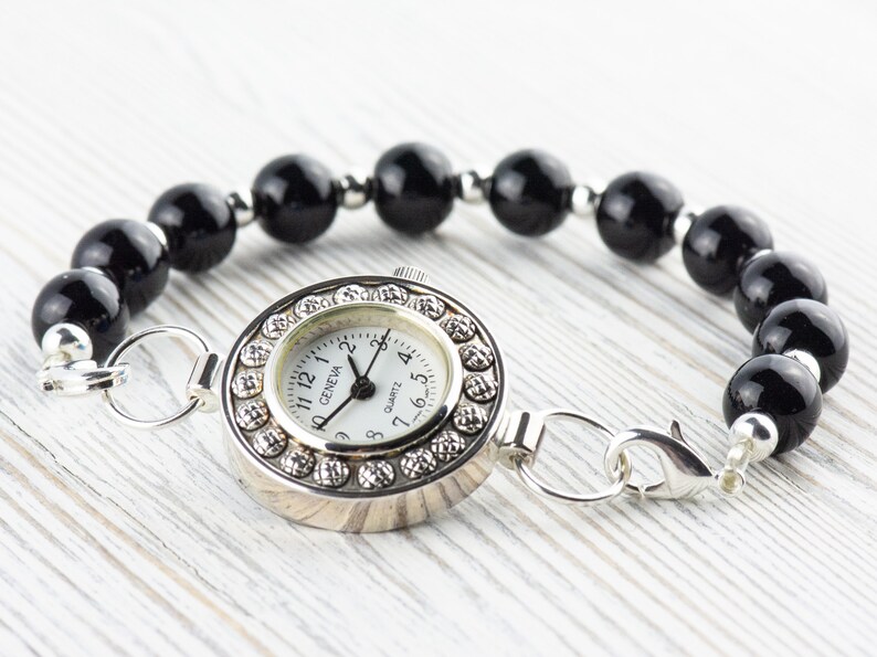 Pu&ograve; includere: Bracciale orologio al quarzo Geneva con quadrante argentato, quadrante bianco e dettagli in cristallo. Il bracciale &egrave; composto da grandi perle nere lucide ed elementi di collegamento argentati.