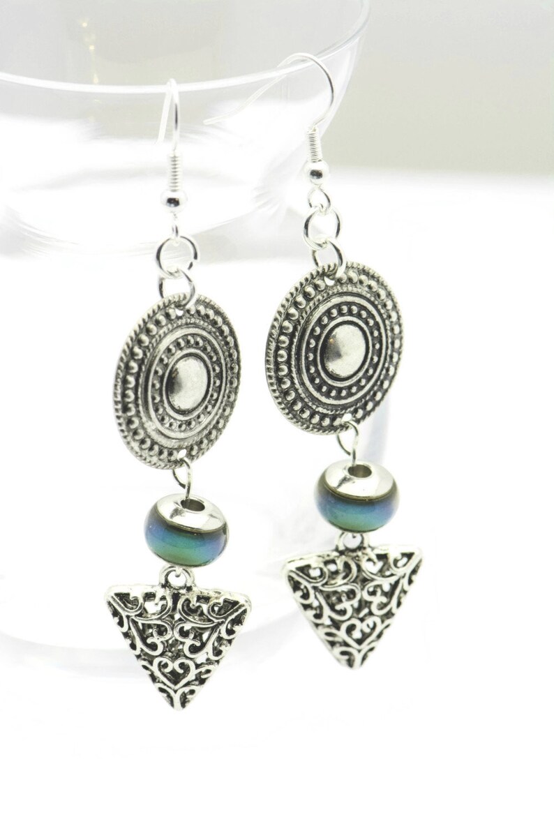 May include: Silver earrings with a round, ornate charm, a blue iridescent bead, and a triangular charm with a filigree design.