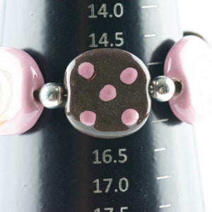 May include: A bracelet made of pink and white ceramic beads with a brown ceramic bead with pink polka dots in the center. The bracelet is on a black measuring tape.
