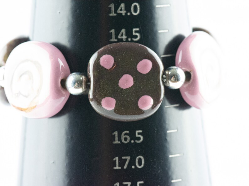 May include: A bracelet made of pink and white ceramic beads with a brown ceramic bead with pink polka dots in the center. The bracelet is on a black measuring tape.