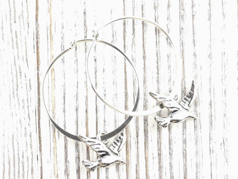 Peut inclure: Boucles d'oreilles cr&eacute;oles argent&eacute;es avec un charm en forme d'oiseau. Les boucles d'oreilles pr&eacute;sentent un design de cr&eacute;oles circulaires avec un pendentif oiseau d&eacute;taill&eacute;. Sur une surface en bois blanc vieilli.