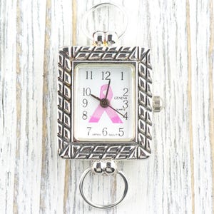 May include: Silver-toned Geneva quartz watch with a rectangular face. The watch face features a pink ribbon design, a symbol of breast cancer awareness. The watch has a decorative, textured bezel and two circular attachments for a chain or strap.