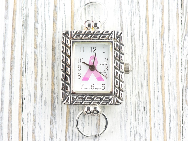May include: Silver-toned Geneva quartz watch with a rectangular face. The watch face features a pink ribbon design, a symbol of breast cancer awareness. The watch has a decorative, textured bezel and two circular attachments for a chain or strap.
