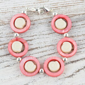 Pu&ograve; includere: Un braccialetto rosa e bianco con una chiusura argentata. Il braccialetto &egrave; fatto di perline rotonde con un centro bianco e un anello esterno rosa.