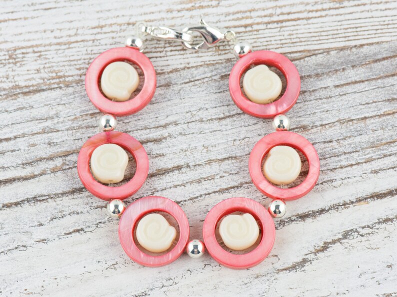 Pu&ograve; includere: Un braccialetto rosa e bianco con una chiusura argentata. Il braccialetto &egrave; fatto di perline rotonde con un centro bianco e un anello esterno rosa.