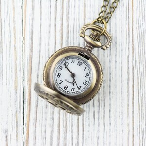 Puede incluir: Un reloj de bolsillo dorado con una esfera blanca y agujas negras. El reloj est&aacute; abierto y la cadena es visible. El reloj est&aacute; sobre una superficie de madera blanca.