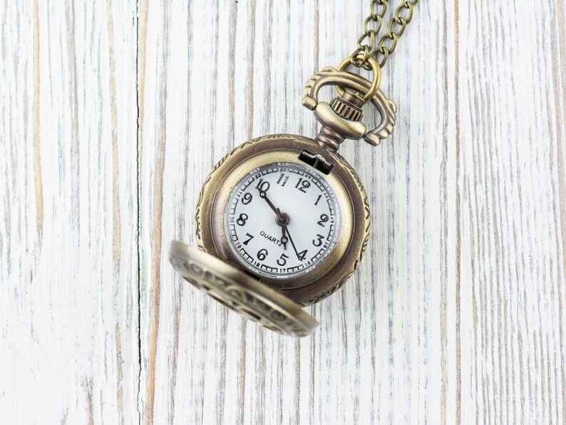 Puede incluir: Un reloj de bolsillo dorado con una esfera blanca y agujas negras. El reloj est&aacute; abierto y la cadena es visible. El reloj est&aacute; sobre una superficie de madera blanca.