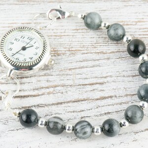 May include: A silver pocket watch with a white face and black hands is attached to a bracelet made of gray and black beads.