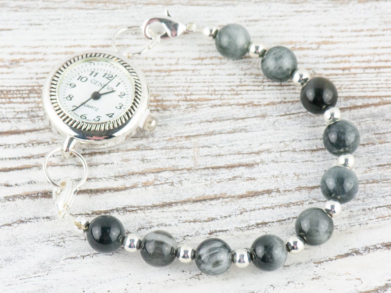 May include: A silver pocket watch with a white face and black hands is attached to a bracelet made of gray and black beads.
