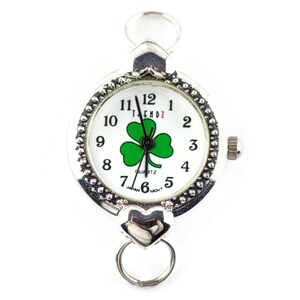 Peut inclure: Montre argent&eacute;e avec un cadran blanc et un motif tr&egrave;fle &agrave; quatre feuilles vert. La montre a un bracelet argent&eacute; avec un fermoir en forme de c&oelig;ur.