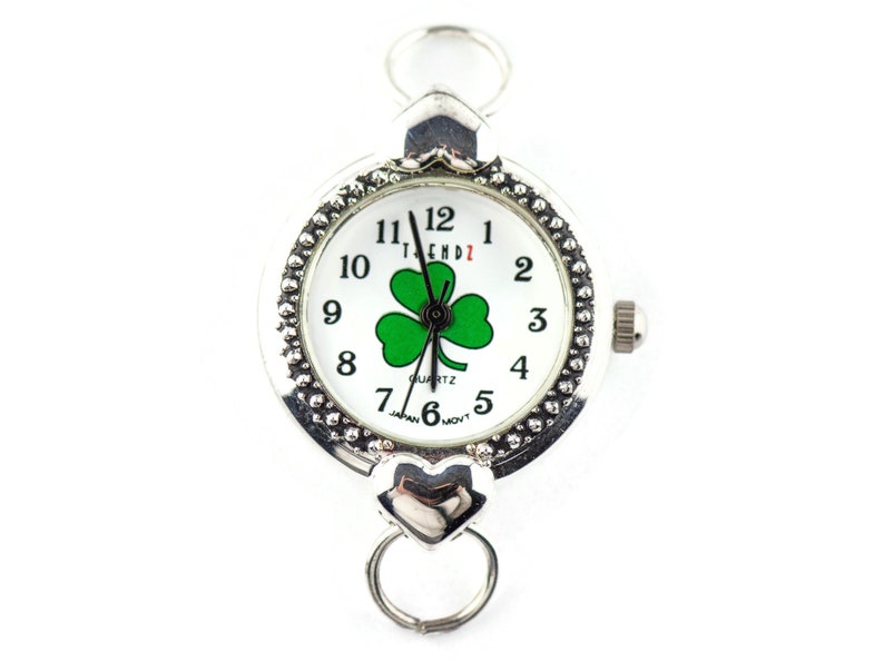 Peut inclure: Montre argent&eacute;e avec un cadran blanc et un motif tr&egrave;fle &agrave; quatre feuilles vert. La montre a un bracelet argent&eacute; avec un fermoir en forme de c&oelig;ur.