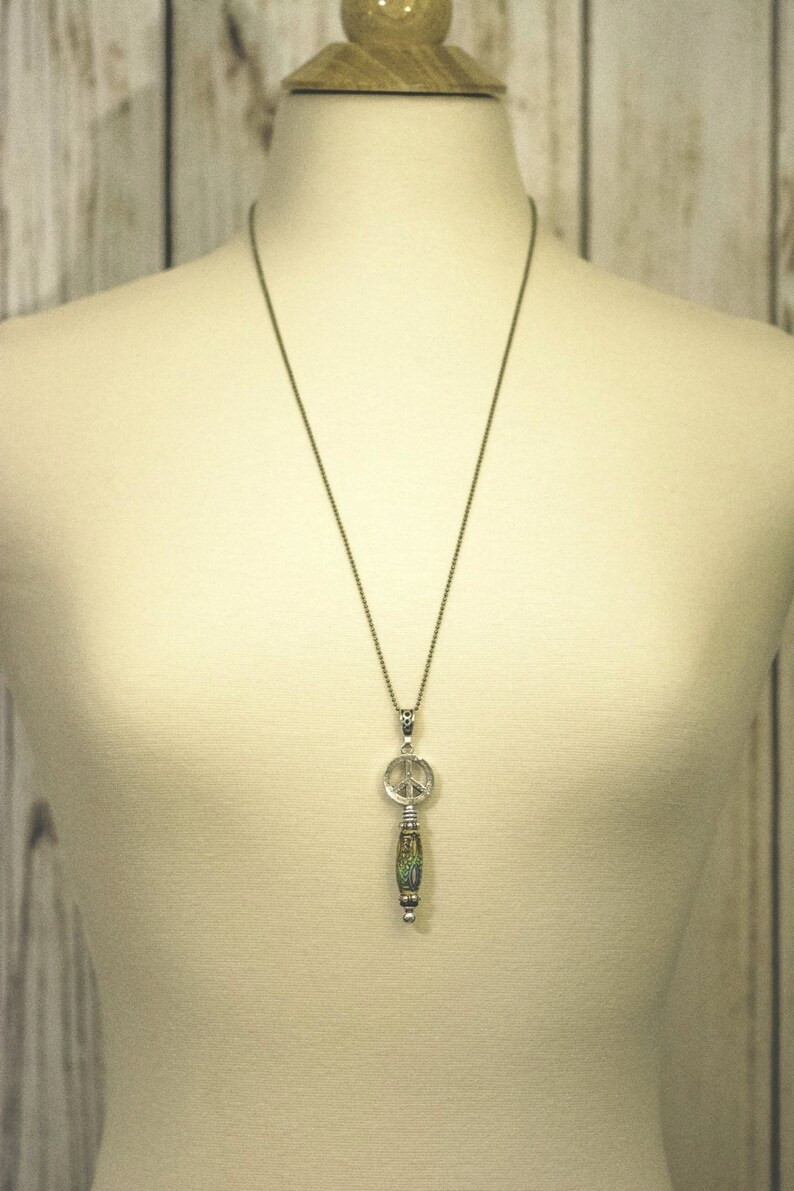 Puede incluir: Un collar con un colgante de plata con el signo de la paz y un abalorio de cuentas verdes y marrones.