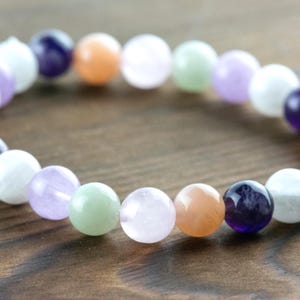 May include: A beaded bracelet featuring a variety of colorful gemstones. The bracelet includes round beads in shades of purple, orange, white, green, and pink. The bracelet is displayed on a wooden surface.