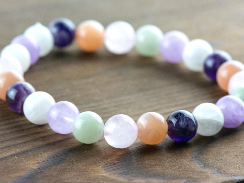 May include: A beaded bracelet featuring a variety of colorful gemstones. The bracelet includes round beads in shades of purple, orange, white, green, and pink. The bracelet is displayed on a wooden surface.