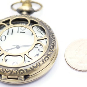 K&ouml;nnte beinhalten: Eine goldfarbene Taschenuhr mit einem verzierten Design und einem wei&szlig;en Zifferblatt. Die Uhr ist etwa so gro&szlig; wie eine deutsche 2-Euro-M&uuml;nze.