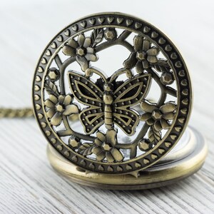 K&ouml;nnte beinhalten: Antike, messingfarbene Taschenuhr mit detailliertem Schmetterlings- und Blumendesign auf dem Deckel. Die Uhr ist ge&ouml;ffnet und zeigt das Zifferblatt. Sie liegt auf einer hellen Holzoberfl&auml;che. Die Uhr hat eine Kette.