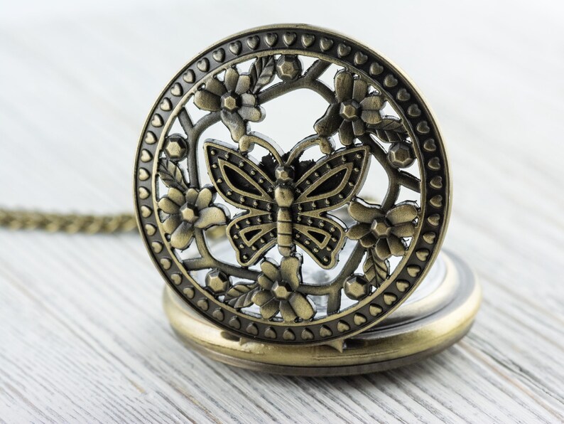 K&ouml;nnte beinhalten: Antike, messingfarbene Taschenuhr mit detailliertem Schmetterlings- und Blumendesign auf dem Deckel. Die Uhr ist ge&ouml;ffnet und zeigt das Zifferblatt. Sie liegt auf einer hellen Holzoberfl&auml;che. Die Uhr hat eine Kette.