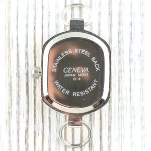 May include: A silver-toned Geneva watch with a stainless steel back, water-resistant design, and a Japanese movement. The watch has a rectangular face with rounded edges and a small dial on the side. It includes a key ring attachment.