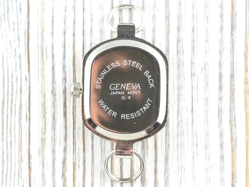 May include: A silver-toned Geneva watch with a stainless steel back, water-resistant design, and a Japanese movement. The watch has a rectangular face with rounded edges and a small dial on the side. It includes a key ring attachment.