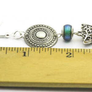 May include: Silver earrings with a round, filigree charm, a blue iridescent bead, and a triangular filigree charm. The earrings are measured against a ruler, showing they are about 1 inch long.