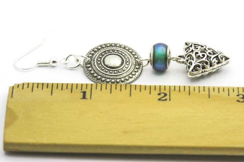 May include: Silver earrings with a round, filigree charm, a blue iridescent bead, and a triangular filigree charm. The earrings are measured against a ruler, showing they are about 1 inch long.