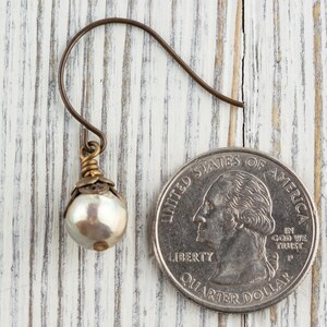May include: A single pearl earring with a bronze-colored hook and decorative accents. The earring features a lustrous, round pearl. The earring is next to a U.S. quarter coin for size comparison. The background is a white-painted wooden surface.