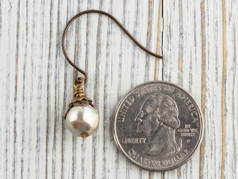 May include: A single pearl earring with a bronze-colored hook and decorative accents. The earring features a lustrous, round pearl. The earring is next to a U.S. quarter coin for size comparison. The background is a white-painted wooden surface.