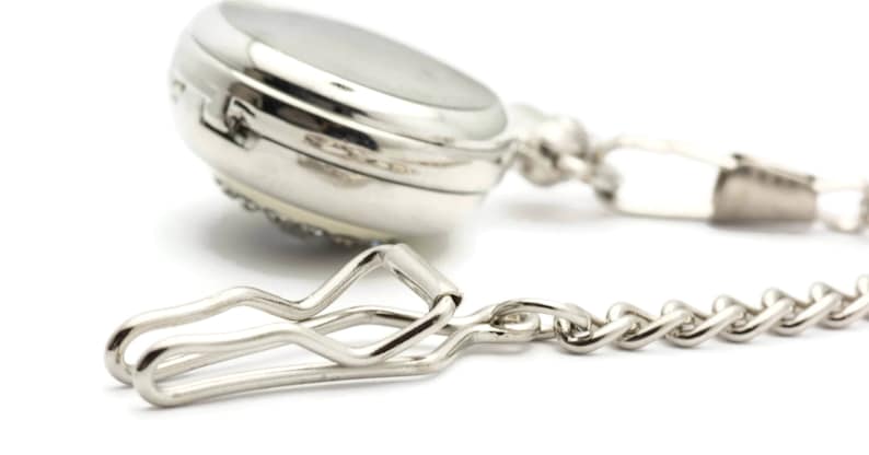 May include: A silver pocket watch with a chain and a clip. The watch has a round face and a silver chain attached to a clip.