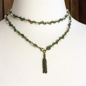 May include: A double-strand necklace with a tassel pendant. The necklace is made of small, linked metal beads in a bronze colour. The beads are interspersed with white and turquoise beads.