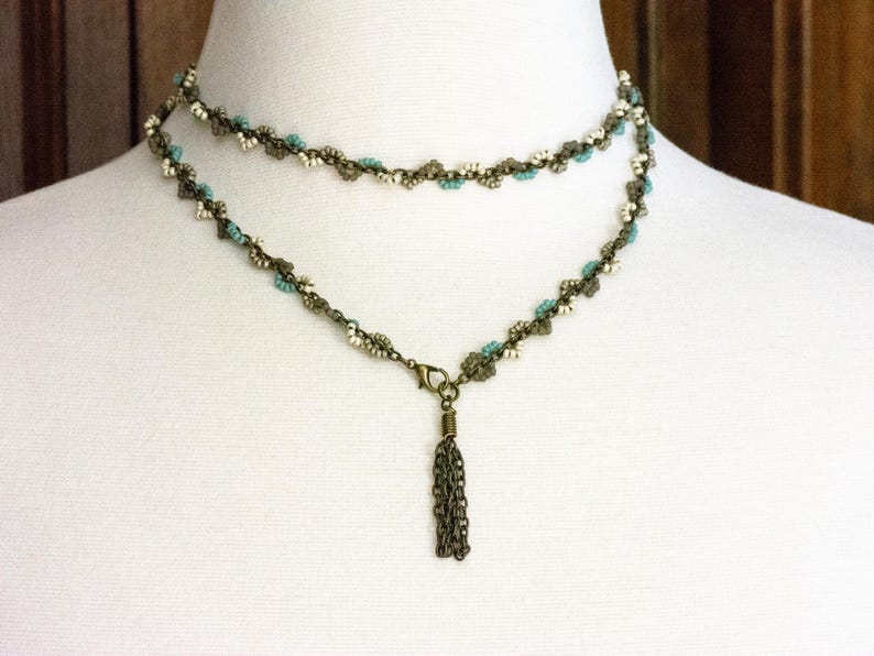 May include: A double-strand necklace with a tassel pendant. The necklace is made of small, linked metal beads in a bronze colour. The beads are interspersed with white and turquoise beads.