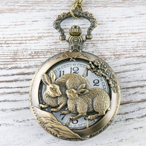 Puede incluir: Reloj de bolsillo antiguo de bronce con un dise&ntilde;o detallado de conejos. La esfera del reloj es blanca con n&uacute;meros y manecillas negras. El reloj est&aacute; suspendido de una cadena a juego. Di&aacute;metro: aprox. 5 cm.