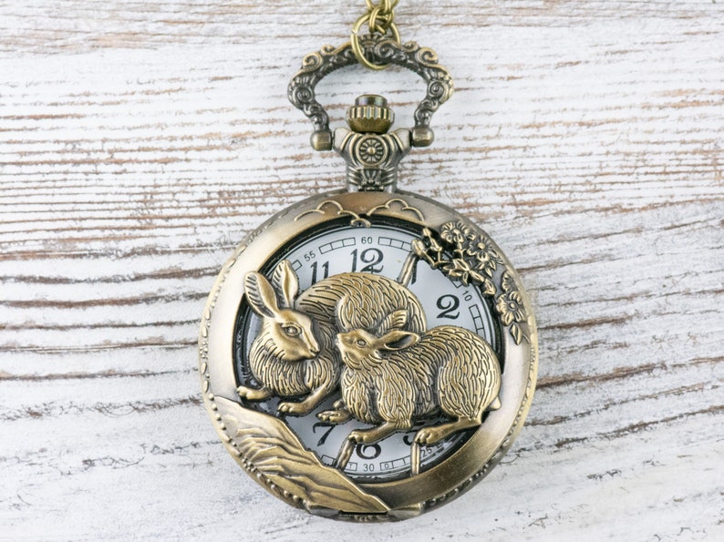 Puede incluir: Reloj de bolsillo antiguo de bronce con un dise&ntilde;o detallado de conejos. La esfera del reloj es blanca con n&uacute;meros y manecillas negras. El reloj est&aacute; suspendido de una cadena a juego. Di&aacute;metro: aprox. 5 cm.